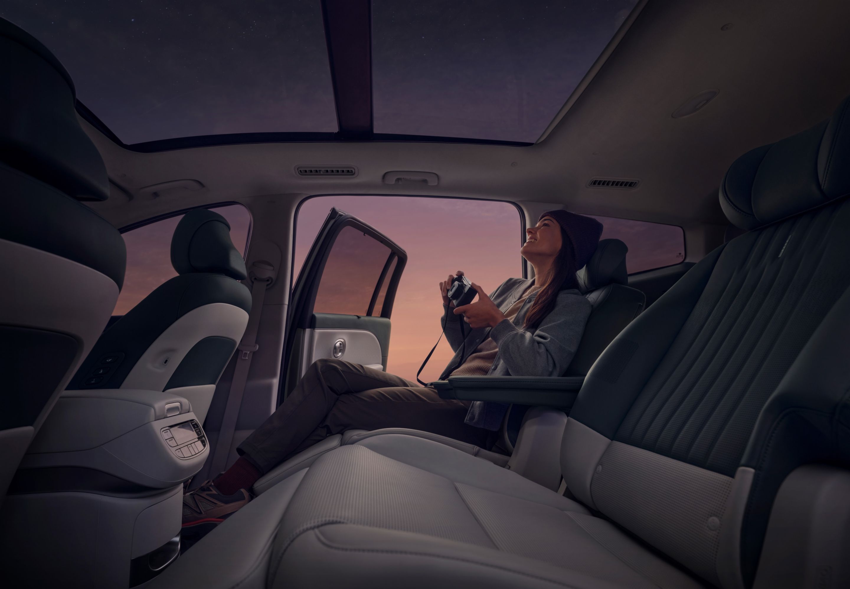 Persona con cámara en el asiento trasero del Hyundai IONIQ 9, puerta abierta hacia un cielo violeta-rosado.